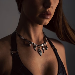 Op de afbeelding: Een zilveren ketting met meerdere bedels, waaronder een schedel, een hand en een hoorn. De ketting wordt gedragen door een persoon met lang bruin haar.