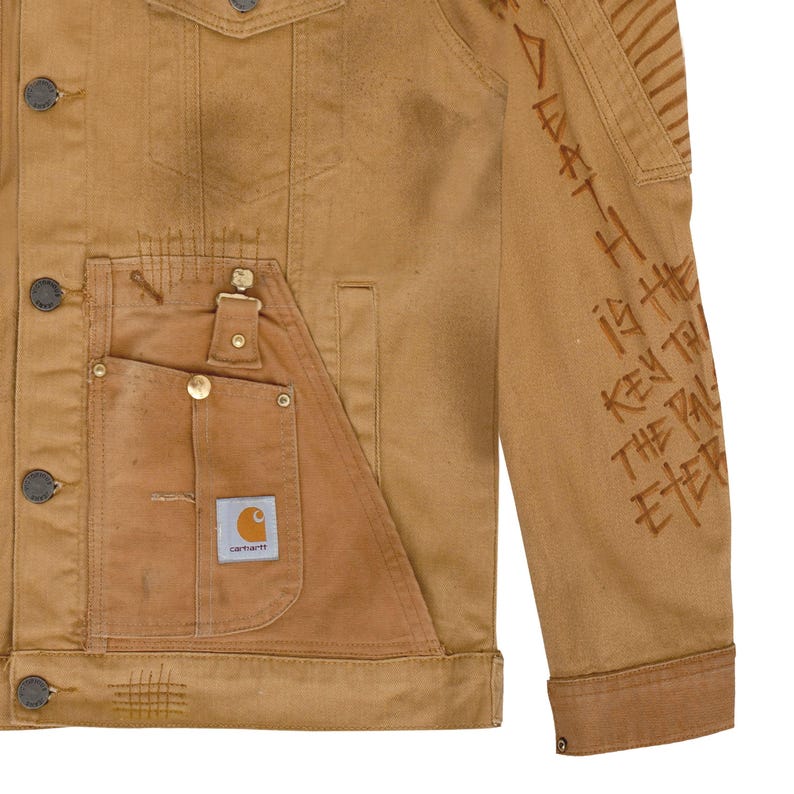 Puede incluir: Chaqueta vaquera Carhartt marr&oacute;n con un dise&ntilde;o &uacute;nico. Cuenta con un bolsillo grande en la parte delantera, botones met&aacute;licos y el logotipo de Carhartt. La manga derecha tiene texto marr&oacute;n: "Death is the key to the eternal".