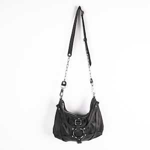 K&ouml;nnte beinhalten: Schwarze Schultertasche aus Leder mit silbernen Ketten- und Ringdetails. Die Tasche hat eine geschwungene Form, einen Rei&szlig;verschluss und einen verstellbaren Riemen. Die Tasche h&auml;ngt vor einem wei&szlig;en Hintergrund.