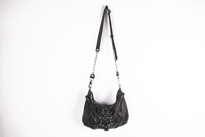 K&ouml;nnte beinhalten: Schwarze Schultertasche aus Leder mit silbernen Ketten- und Ringdetails. Die Tasche hat eine geschwungene Form, einen Rei&szlig;verschluss und einen verstellbaren Riemen. Die Tasche h&auml;ngt vor einem wei&szlig;en Hintergrund.