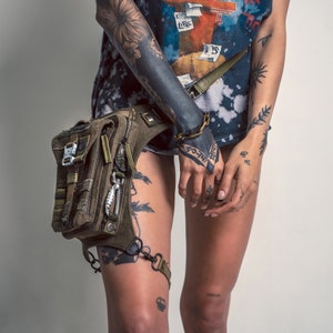 May include: A person with tattoos wearing a blue and white shirt with a graphic print and a green and brown tactical leg bag with straps.