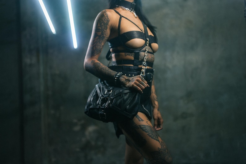 K&ouml;nnte beinhalten: Schwarzes Leder-Harness- und Rock-Set mit passender Tasche. Das Harness hat ein Kettendetail und die Tasche einen Schnallenverschluss. Das Outfit wird mit einem Halsband und Armband getragen. Die Person hat T&auml;towierungen an Armen und Beinen.