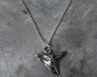 May include: A silver-colored pendant necklace featuring a shark tooth design. The pendant is intricately detailed and hangs from a delicate chain. The necklace is displayed against a gray background.