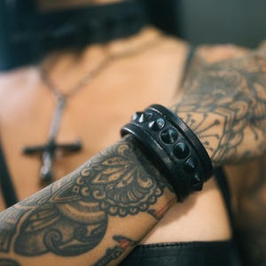 以下が含まれることがあります： 銀のスタッズが付いた黒い革のブレスレット。ブレスレットはタトゥーのある腕につけられています。