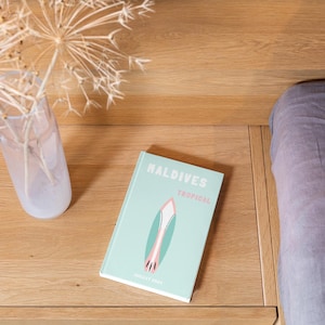 Peut inclure: Un livre bleu clair avec le titre "Maldives Tropical" et une illustration de planche de surf rose et verte. Le livre est posé sur une surface en bois à côté d'un vase en verre transparent avec des fleurs séchées.