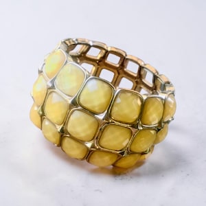 May include: A gold-toned bracelet with a grid-like design and yellow, square-shaped stones. The bracelet is made of metal and features a stretchable design. The stones are a pale yellow color and are set in a grid pattern.