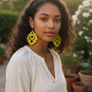 May include: A woman wearing vibrant yellow, openwork chandelier earrings. The earrings feature an intricate, symmetrical pattern. She is wearing a white, long-sleeved blouse with a V-neck. The background is a blurred outdoor scene with greenery and plant pots.
