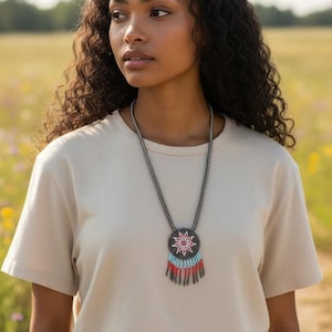 Può includere: Una donna indossa una maglietta beige e jeans blu, con una lunga collana di perline. La collana presenta un ciondolo circolare con un motivo floreale rosa, rosso e nero, e frange blu e rosse.