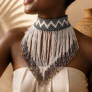 May include: A beaded choker necklace featuring a wide black and white zigzag pattern band. Long, vertical white beaded fringe strands extend downwards from the band, with the lower edges accented by small, multi-coloured beads in blue, red, and yellow.