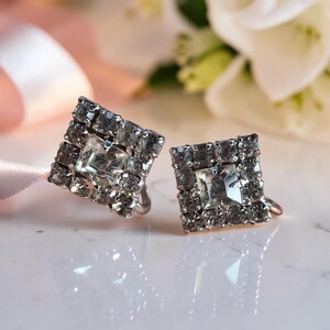 May include: A pair of square-shaped clip-on earrings featuring a central clear square-cut stone surrounded by a border of smaller, round, faceted clear crystals set in a silver-toned metal frame, reflecting light on a smooth surface.
