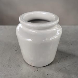 May include: A small, white ceramic jar with a rounded body and a wide, slightly flared rim. The jar has a smooth, glossy finish and appears to be empty. The background is a neutral gray, highlighting the jar's simple design.