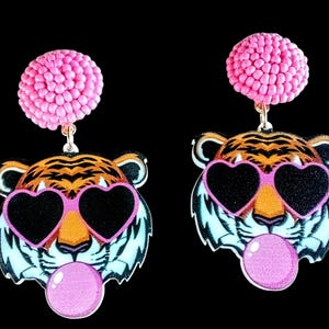 May include: Pink beaded dangle earrings with a tiger face charm wearing pink heart-shaped sunglasses and blowing a pink bubble.