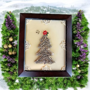 May include: A framed Christmas tree art piece. The tree is made of silver-colored material and topped with a red poinsettia. The frame is dark brown, and the background is a light beige color. Decorative elements include snowflakes and a small bird.