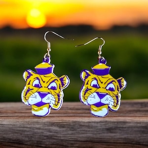 May include: A pair of yellow and purple tiger-faced earrings with gold-colored hooks. The earrings feature a cartoon-like design with a white outline. The background shows a sunset over a green field.