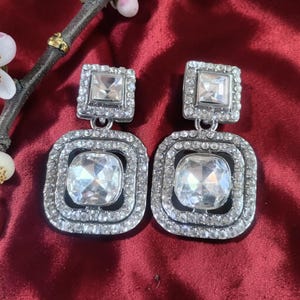 May include: A pair of silver-toned, square drop earrings with large, clear, faceted crystals. Each earring features a smaller square crystal at the top and a larger, square crystal at the bottom, surrounded by smaller crystals. The earrings are displayed on a red surface.