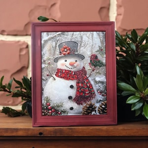 May include: A framed winter scene depicts a snowman with a top hat, red scarf, and black buttons. A red bird sits on a branch. The frame is a distressed red colour, and the background shows a snowy forest.