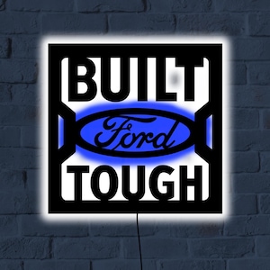 May include: Black and white square sign with the words "BUILT TOUGH" and the Ford logo in blue. The sign is illuminated with a white glow and has a black border. The sign is mounted on a brick wall.