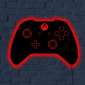 May include: Black Xbox controller-shaped wall art with a red neon outline and internal details. The controller features a red Xbox logo, buttons, and directional pad. The art is mounted on a dark blue brick wall.