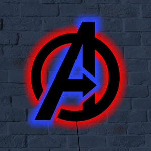 Puede incluir: Logotipo negro de los Vengadores con un resplandor de neón rojo y azul, montado en una pared de ladrillos grises. El logotipo presenta una gran letra "A" con una flecha, rodeada por un círculo. Se ve un cable de alimentación negro.