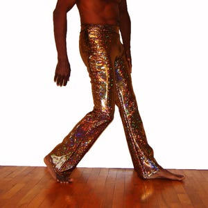 Op de afbeelding: Gouden, glinsterende broek met een holografisch effect. De broek heeft een uitlopende pijp en een hoge taille. De stof reflecteert meerdere kleuren, waardoor een disco-achtige uitstraling ontstaat. De broek wordt getoond op een persoon die op een houten vloer loopt.
