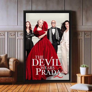 May include: A framed movie poster for "The Devil Wears Prada 2" featuring the cast. The poster has a black frame and is displayed on a wooden floor. The title is in white, with a red devil's tail.