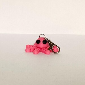 May include: A pink octopus charm with black eyes and a silver-colored clasp. The octopus has eight tentacles and is made of a smooth material. The charm is designed to be attached to a keychain or bag.