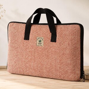 May include: A rectangular laptop sleeve featuring a red and white herringbone pattern with black fabric handles and trim. A central rectangular patch displays the text Himalayan Hemp THC Free Made in Nepal with a green cannabis leaf graphic icon.
