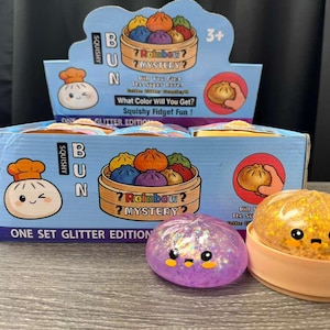 May include: A box set of Squishy Bun toys, featuring a glitter edition. The box is blue with cartoon illustrations of buns and text that reads "? Rainbow Mystery?". Two squishy toys are visible: one purple and one orange, both with cute faces.