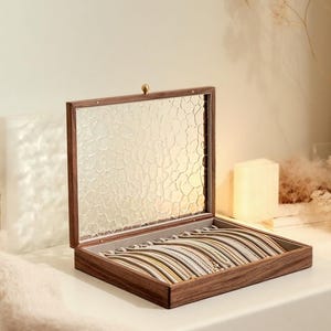 Könnte beinhalten: Eine Holzschmuckschatulle mit strukturiertem Glasdeckel, geöffnet, um eine Reihe von Armbändern in verschiedenen Farben zu zeigen. Die Schatulle ist aus dunklem Holz gefertigt und hat einen messingfarbenen Verschluss. Die Armbänder sind ordentlich angeordnet.