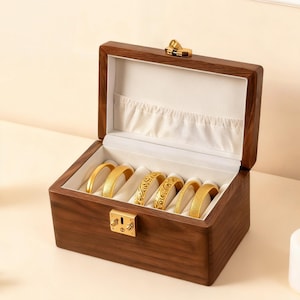 Peut inclure: Une boîte à bijoux en bois avec un fermoir doré, ouverte pour révéler plusieurs bracelets dorés. L'intérieur est doublé de tissu blanc. La boîte est sur une surface claire, avec une bougie blanche allumée en arrière-plan.