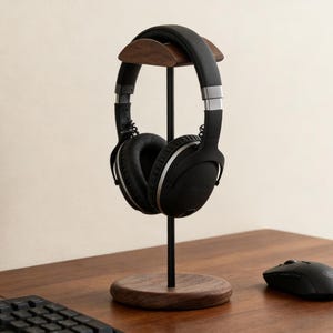 Könnte beinhalten: Ein Kopfhörerständer aus Holz mit einem schwarzen Metallpfosten und einem runden Sockel. Schwarze Kopfhörer mit silbernen Akzenten liegen auf dem Ständer. Eine schwarze Computermaus befindet sich auf dem Holzschreibtisch.