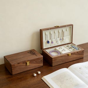 Könnte beinhalten: Zwei hölzerne Schmuckkästchen, eines offen und eines geschlossen, stehen auf einer Holzoberfläche. Die offene Box zeigt Halsketten, Ringe und ein Armband. Die geschlossene Box hat ein Blumendesign. Ein Buch und Perlenohrringe befinden sich im Vordergrund.