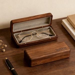 Könnte beinhalten: Geöffnete Brillenetui aus Holz mit einer runden, goldgerahmten Brille. Das Etui ist mit grauem Stoff ausgekleidet. Ein geschlossenes Holzgehäuse mit dem Namen "Sabine" daneben. Ein schwarzer Stift und Bücher sind ebenfalls sichtbar.