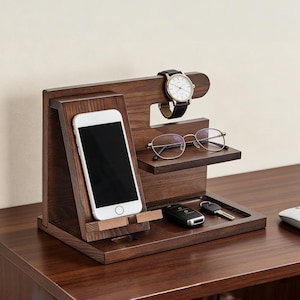 Könnte beinhalten: Ein dunkelbrauner Schreibtisch-Organizer aus Holz mit Smartphone, Brille, Uhr und Schlüsseln. Die Uhr hat ein schwarzes Lederarmband und ein weißes Zifferblatt. Der Organizer hat einen Schlitz für das Telefon und ein kleines Tablett für Schlüssel.