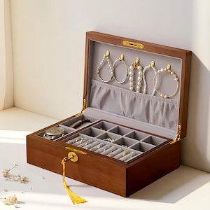 Könnte beinhalten: Eine offene Schmuckschatulle aus Holz mit Fächern und einem grauen Samtfutter. Die Schatulle enthält verschiedene Schmuckstücke, darunter Armbänder, Ringe und eine Uhr. Die Schatulle ist braun mit goldfarbener Hardware und einer gelben Quaste.