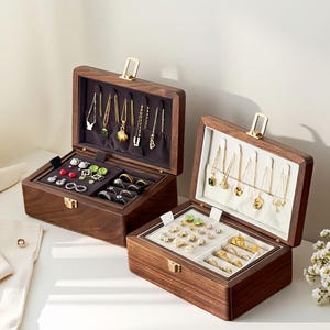 Könnte beinhalten: Zwei offene Schmuckkästchen aus Holz mit verschiedenen Halsketten, Ringen und Ohrringen. Die Kästchen sind dunkelbraun mit goldfarbenen Verschlüssen und Scharnieren. Die Innenräume sind mit dunkelgrauem und weißem Stoff ausgekleidet.