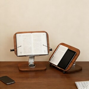 May include: Two wooden book stands with adjustable metal supports. One holds an open book, the other a notebook and tablet. The stands have a dark wood finish and are on a wooden surface.