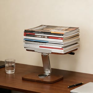 May include: A wooden magazine rack with a silver metal support, holding a stack of magazines. The rack is on a wooden desk with a glass of water, a pen, and a laptop.