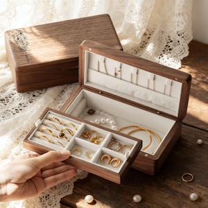 Könnte beinhalten: Eine offene Holzschmuckschatulle mit weiß ausgekleidetem Innenraum und Fächern für Ringe, Halsketten und Armbänder. Die Schatulle ist mit Goldschmuck gefüllt. Der Deckel hat ein Blumendesign und das Wort "Alice".