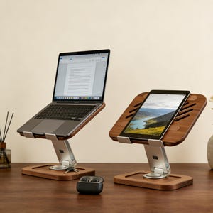 May include: Two wooden laptop and tablet stands with silver metal supports. A laptop displays a document, and a tablet shows a landscape image. Both stands are on a wooden surface. A pair of black wireless earbuds sits on the table.