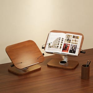 May include: Two wooden book stands with adjustable angles sit on a dark brown desk. One stand holds an open book with images of buildings and people. The stands have a light brown base and a darker brown, angled top.