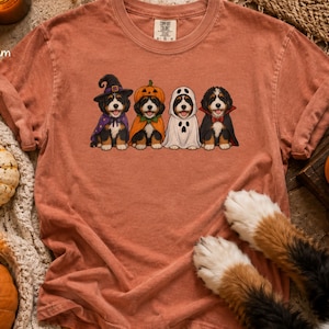 May include: A terracotta-colored short-sleeve t-shirt featuring a graphic of four Bernese Mountain Dog puppies dressed in Halloween costumes: a witch, a pumpkin, a ghost, and a vampire. The shirt has a crew neckline and a Comfort Colors label.