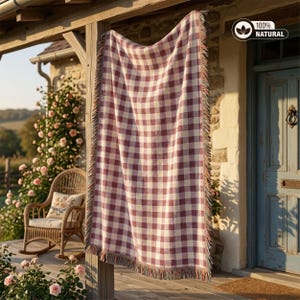 Può includere: Una coperta rettangolare a quadri rossi e bianchi, con frange, appesa su un portico. La coperta è esposta all'aperto, con una sedia a dondolo in vimini e rose visibili. L'etichetta mostra "100% Natural".