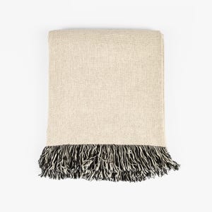 May include: A rectangular throw blanket featuring a solid light beige woven fabric body. The bottom edge is finished with a dense, long fringe composed of alternating black and white threads, creating a high-contrast textured border.