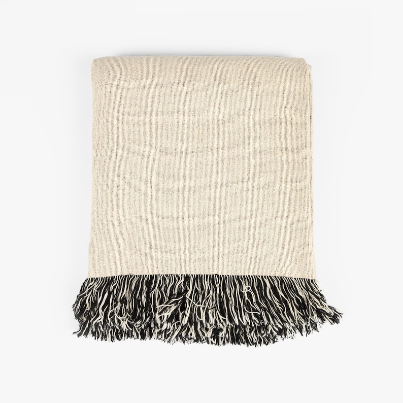 May include: A rectangular throw blanket featuring a solid light beige woven fabric body. The bottom edge is finished with a dense, long fringe composed of alternating black and white threads, creating a high-contrast textured border.