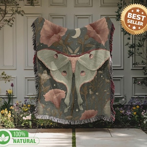 Könnte beinhalten: Eine gewebte Decke mit einem Nachtfalter- und Blumendesign in Grün, Rosa und Grau. Die Decke hat einen Fransenrand und ist mit "100% NATURAL" gekennzeichnet. Das Design beinhaltet einen Mond und Sterne.