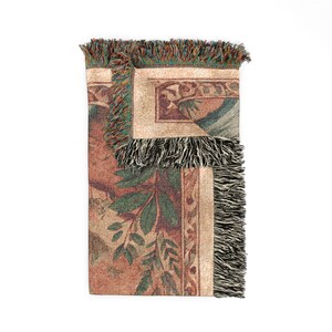 May include: A woven throw blanket featuring a folded corner that reveals a multi-colored design with green leaves, brown tones, and decorative borders. The edges of the blanket are finished with a textured, multi-colored fringe trim.