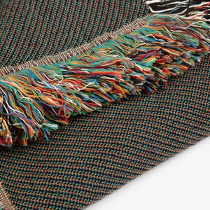 May include: A woven textile blanket featuring a dark, multi-colored micro-check pattern of black, green, and orange threads. The edge of the blanket is finished with a thick, decorative fringe composed of vibrant, multi-colored yarn strands.