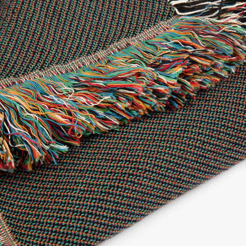 May include: A woven textile blanket featuring a dark, multi-colored micro-check pattern of black, green, and orange threads. The edge of the blanket is finished with a thick, decorative fringe composed of vibrant, multi-colored yarn strands.