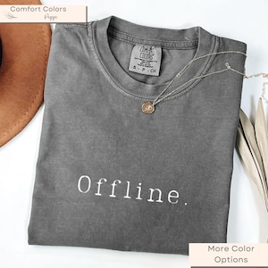Könnte beinhalten: Ein graues T-Shirt mit dem Wort "Offline" in weißer Schrift. Das Shirt hat einen Rundhalsausschnitt und kurze Ärmel. Eine silberne Halskette mit einem runden Anhänger liegt auf dem Shirt. Das Shirt ist von der Marke "Comfort Colors".